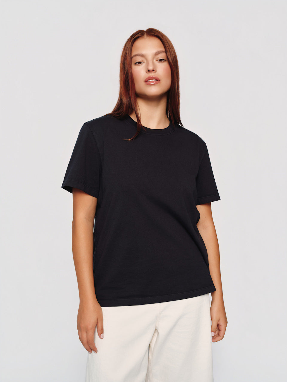A woman modeling a black short-sleeve crewneck t-shirt paired with off-white trousers, shot against a solid light gray studio background.