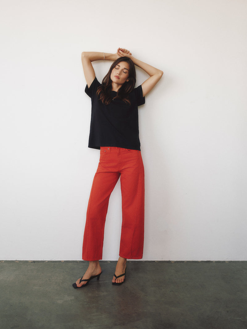 A woman posing in a black short-sleeve t-shirt and bright red wide-leg cropped pants with black strappy heels, standing against a plain white wall.
