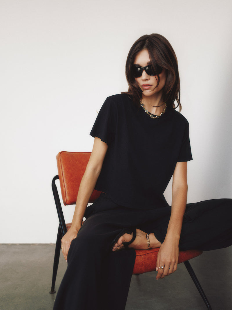 A woman wearing a black short-sleeve top and black wide-leg pants sits on an orange chair against a plain white wall.