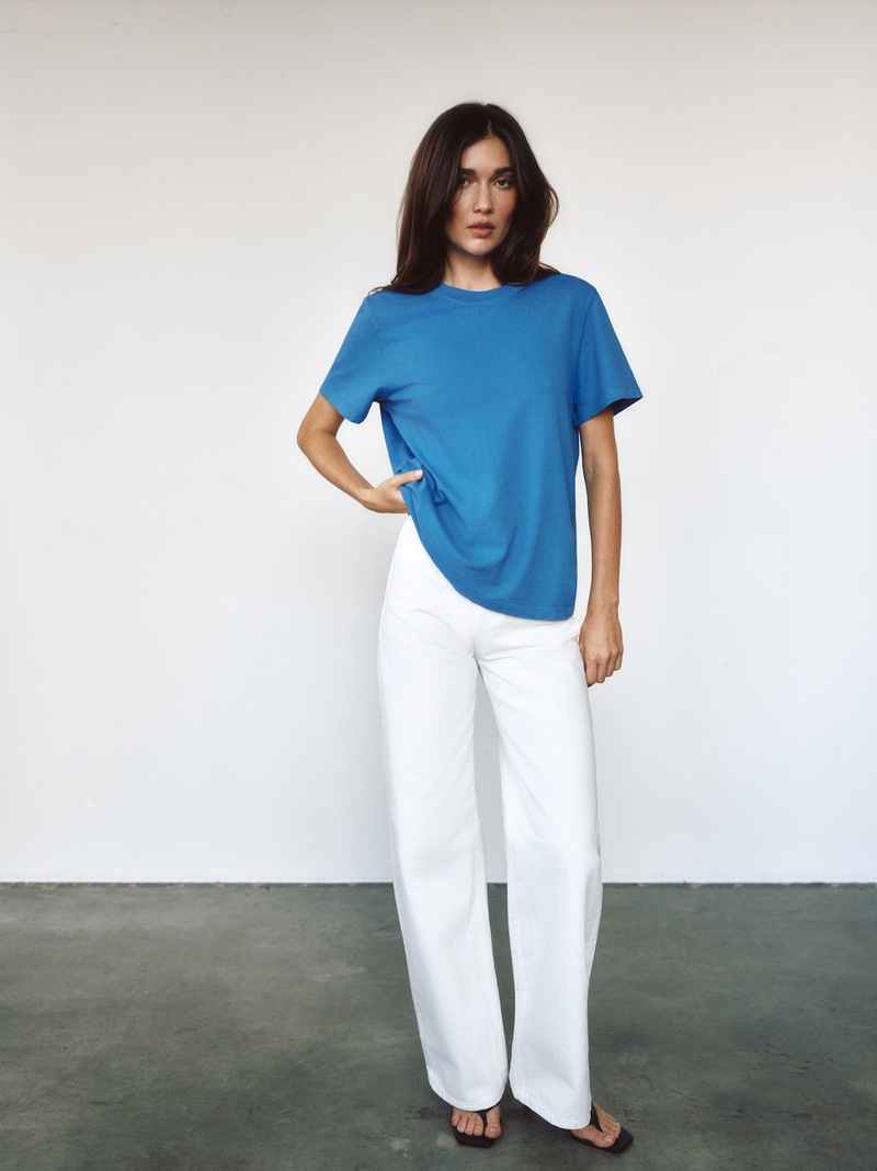 A model wearing a short-sleeve blue t-shirt and white wide-leg trousers, standing against a plain white wall with a gray floor.