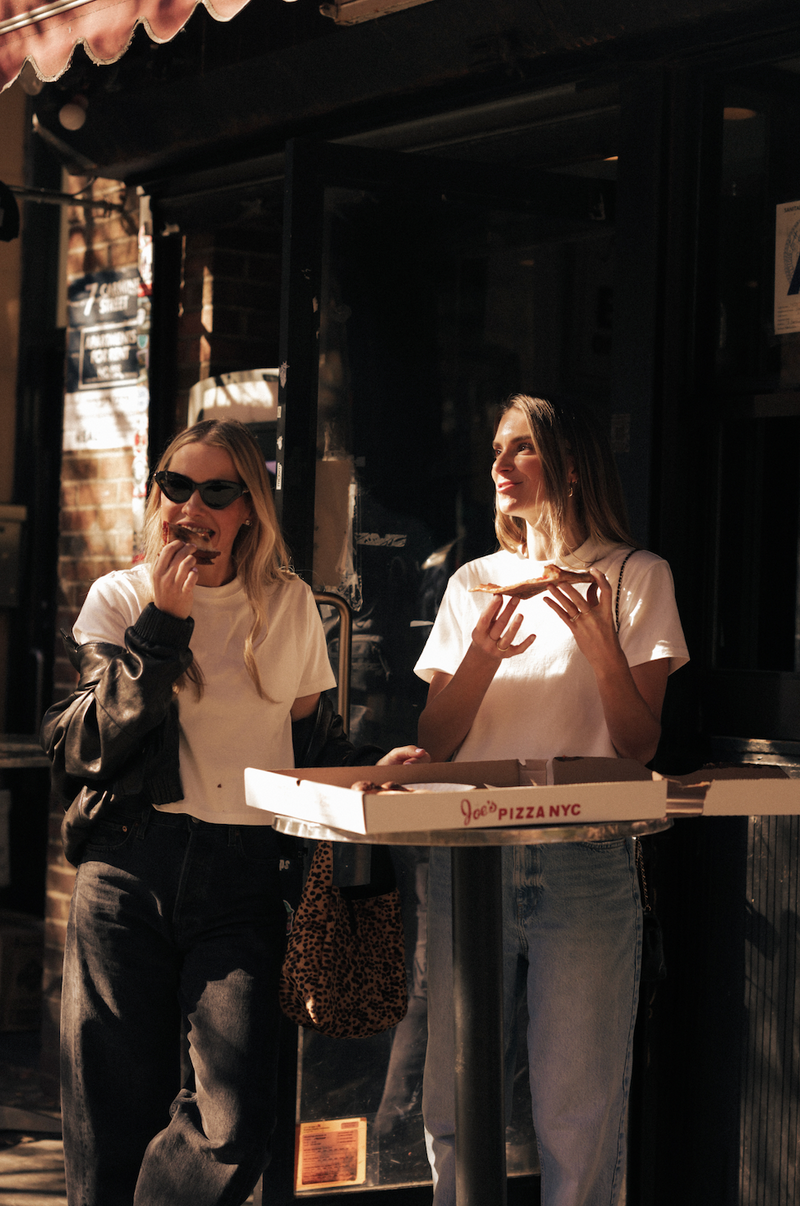 Two women wearing Saint Sirène white t-shirts while sharing pizza outdoors.